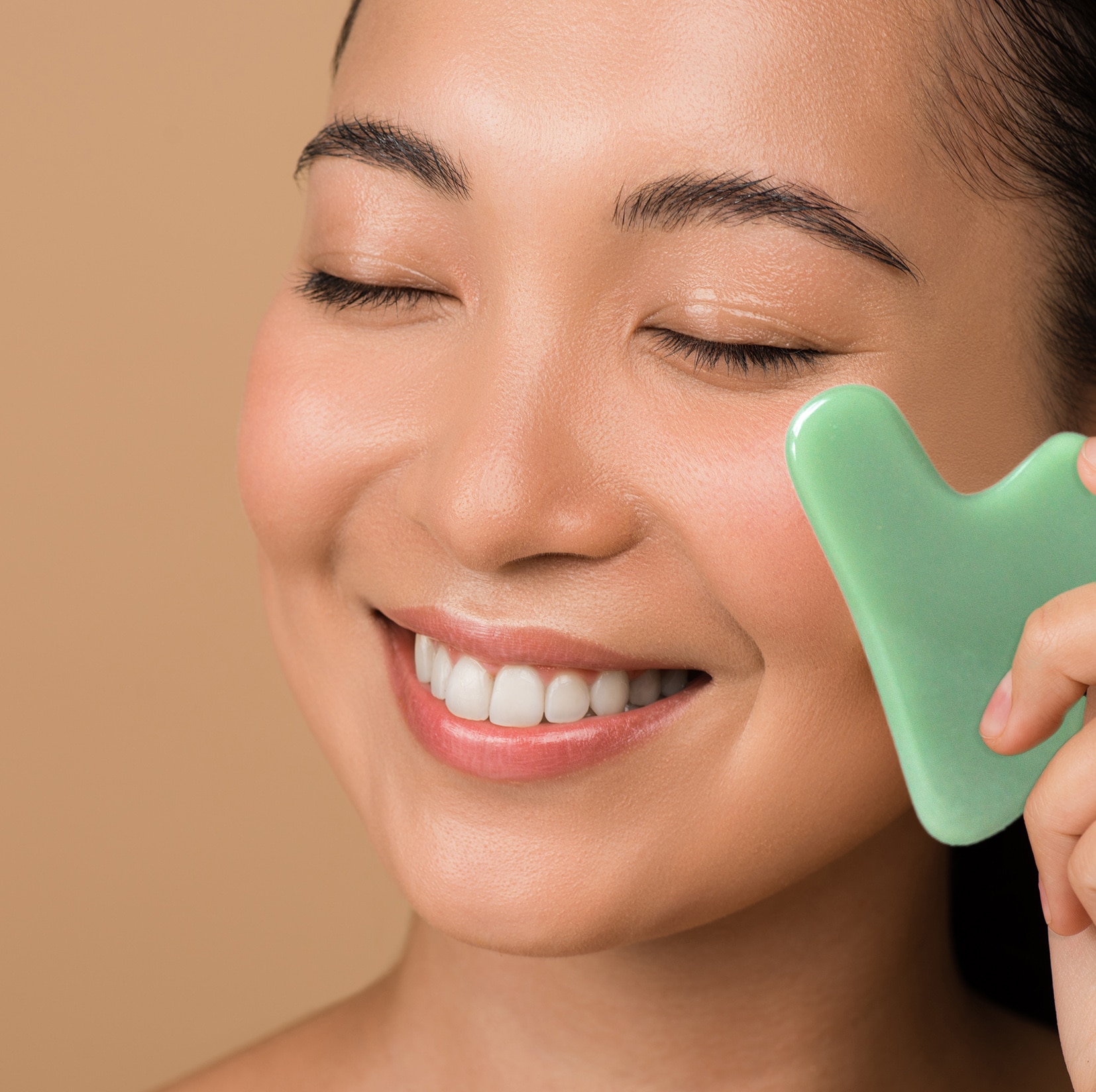 Smiling Beautiful Naked Asian Girl With Closed Eyes Using Facial Jade Gua Sha - Best Natural anti-aging and relaxation